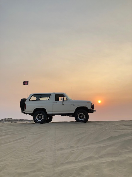 1981 Ford Bronco - A white 1980 Ford Bronco is parked on a sand dune during sunset, with a pink ribbon flag flying from a pole.