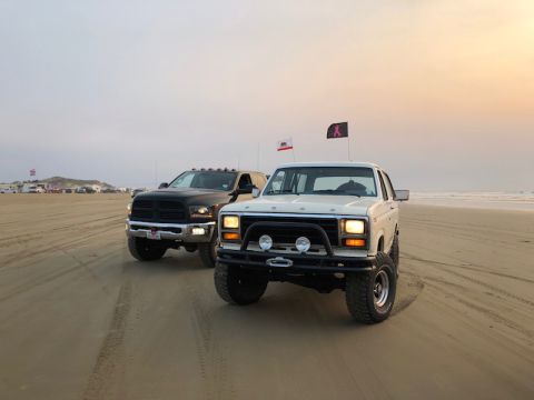1981 Ford Bronco - A white 1981 Ford Bronco and a black Dodge Ram truck are parked on a sandy beach with the ocean in the background.