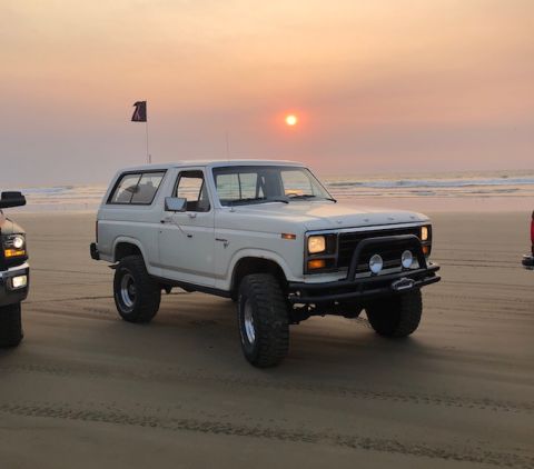 1981 Ford Bronco - A white Ford Bronco sits on the sand near the ocean waves as the sun sets.