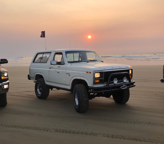 white Ford Bronco parked on a beach with the sun setting over the ocean