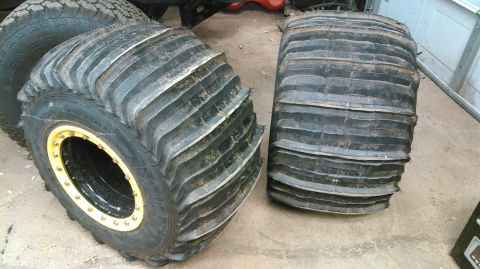 1978 Ford Bronco - Two large, aggressive off-road tires are shown on a concrete floor. The tire on the left is mounted on a black rim with a yellow beadlock,…