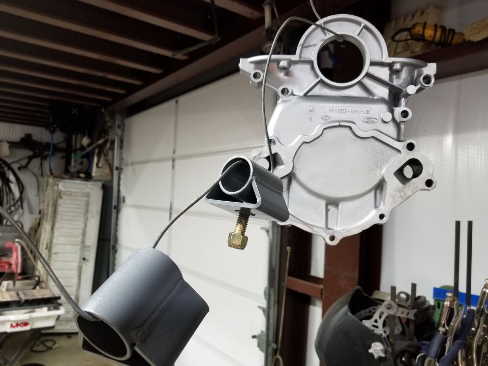 Aluminum engine timing cover with part number SF-F8TE-6059-JB hanging, attached to a metal bracket