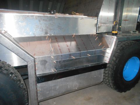1992 Ford Bronco - A close-up view of the unfinished aluminum body of a custom vehicle, showing riveted panels and a blue wheel rim. The vehicle appears to be…