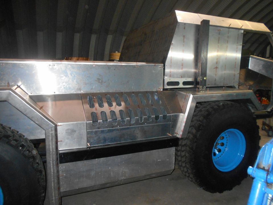 Custom vehicle with aluminum body, large tires, and blue rims in a workshop.