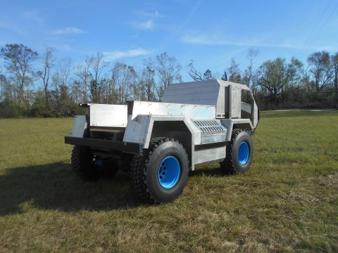 1992 Ford Bronco - A custom-built, four-wheeled vehicle with a light gray, angular body and bright blue wheels sits in a grassy field under a blue sky.