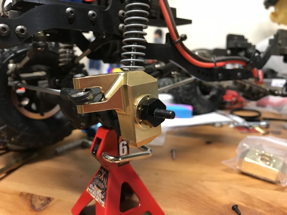 Brass steering knuckle assembly on a red jack stand, RC vehicle suspension in background.