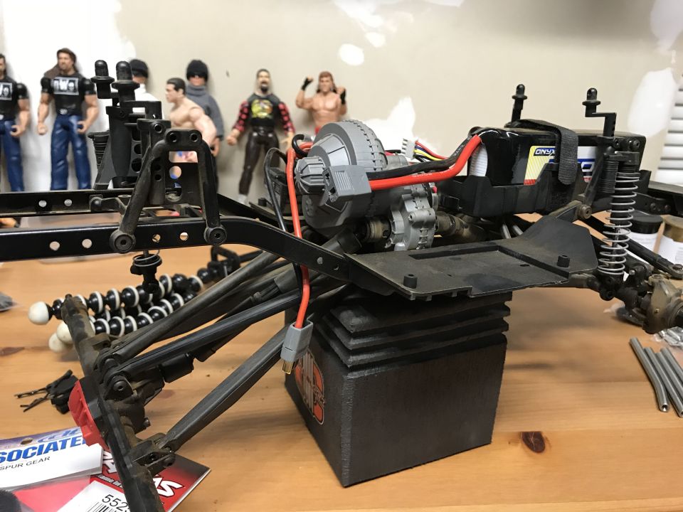 RC car chassis with battery and motor, action figures in background.