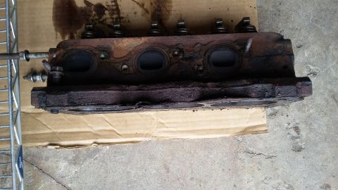 Corroded engine cylinder head with visible valves and ports, resting on cardboard.