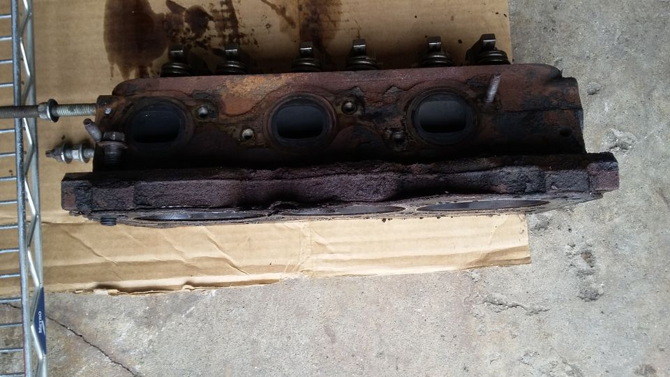 Corroded engine cylinder head with visible valves and ports, resting on cardboard.