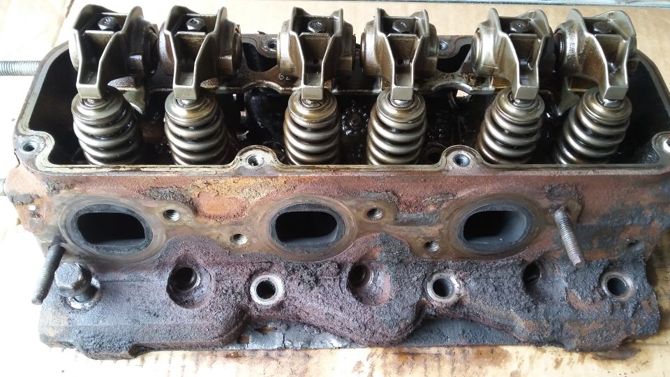 Used V6 engine cylinder head with rocker arms, valve springs, and ports, covered in oil and grime.
