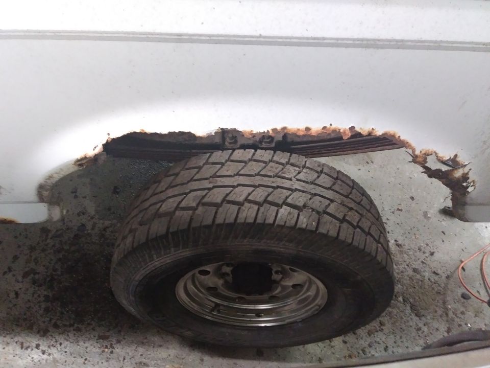 Heavily rusted wheel well opening on a white vehicle, showing leaf springs and a tire.