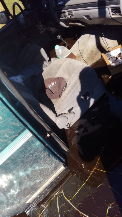 1996 Ford Bronco - Interior view of a flooded vehicle showing the driver's seat, dashboard, and gear shifter. A brown baseball cap with a Ford logo rests on t…