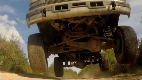 1996 Ford Bronco - The underside of a lifted 1996 Ford Bronco is captured mid-air as it jumps over a dirt path. The vehicle's suspension and large off-road ti…