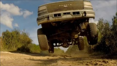 1996 Ford Bronco - A lifted 1996 Ford Bronco is captured mid-air, jumping over a dirt track with trees and a cloudy sky in the background. The underside of th…