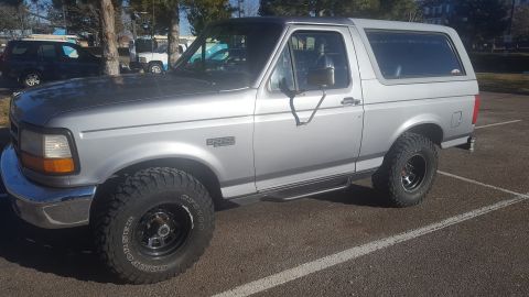 1995 Ford Bronco - A silver 1995 Ford Bronco is parked in a lot, showing its side profile. The vehicle has aftermarket tires and black rims, with running boar…