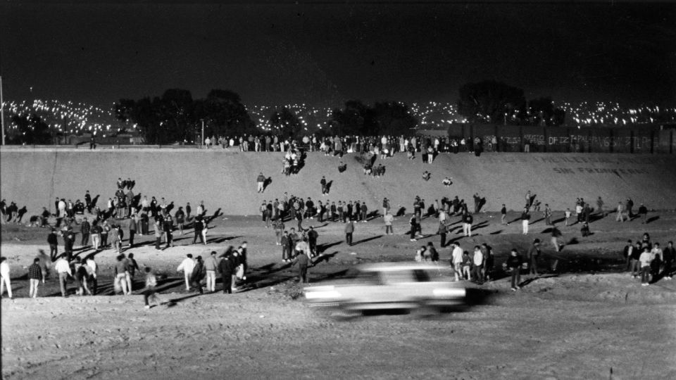 Blurry white car driving past a crowd at night with city lights in the background