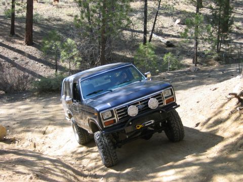 1985 Ford Bronco - 1985 Ford Bronco navigates a steep, dusty off-road path with trees in the background.