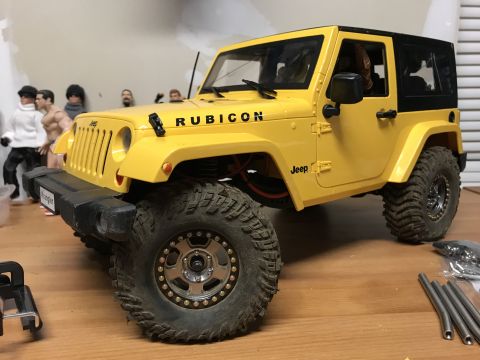 1989 Ford Bronco - A yellow toy Jeep Wrangler Rubicon is shown from a 3/4 front angle, with other toy figures in the background.
