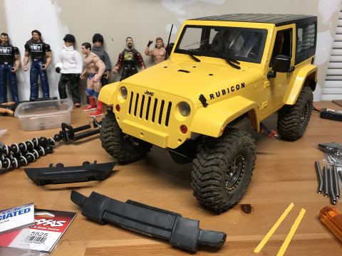 1989 Ford Bronco - RC Jeep Rubicon model with toy figures and unassembled parts on a workbench.
