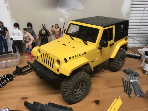 1989 Ford Bronco - A yellow Jeep Rubicon model sits on a wooden surface, surrounded by action figures and hobby supplies.