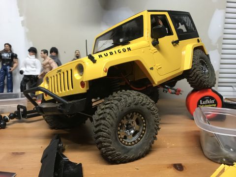 1989 Ford Bronco - A yellow remote-controlled Jeep Wrangler is positioned on a workbench, with several action figures visible behind it.