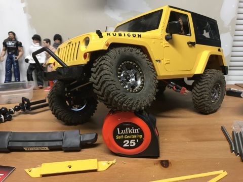 1989 Ford Bronco - A yellow toy Jeep Wrangler Rubicon is elevated on a tape measure, with action figures in the background.