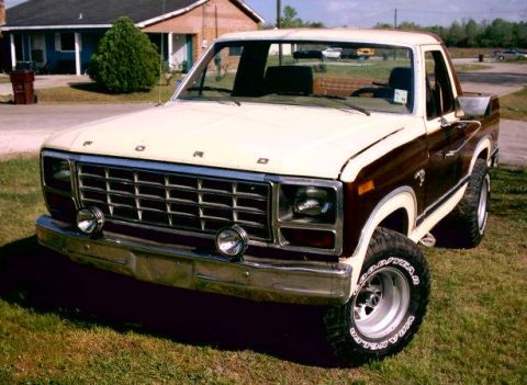 1980 Ford Bronco - lifted two-tone brown and tan Ford 4x4 pickup with FORD-lettered grille and Goodyear tires parked on a lawn.