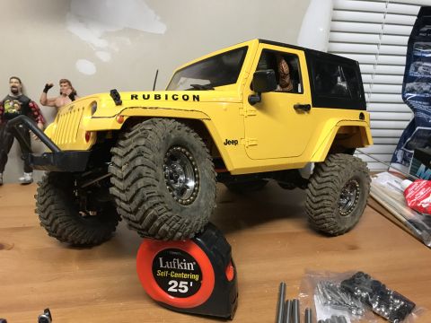 1989 Ford Bronco - A yellow Jeep Rubicon model is displayed on a table, resting on a measuring tape, with various parts and figures in the background.