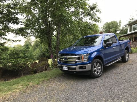 2018 Ford F-150 - A blue 2018 Ford F-150 is parked on a gravel driveway in front of a wooded area and a house.