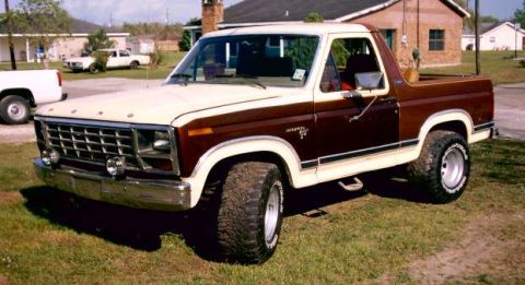 1980 Ford Bronco - A two-tone 1980 Ford Bronco is parked on a grassy area with a white pickup truck in the background.
