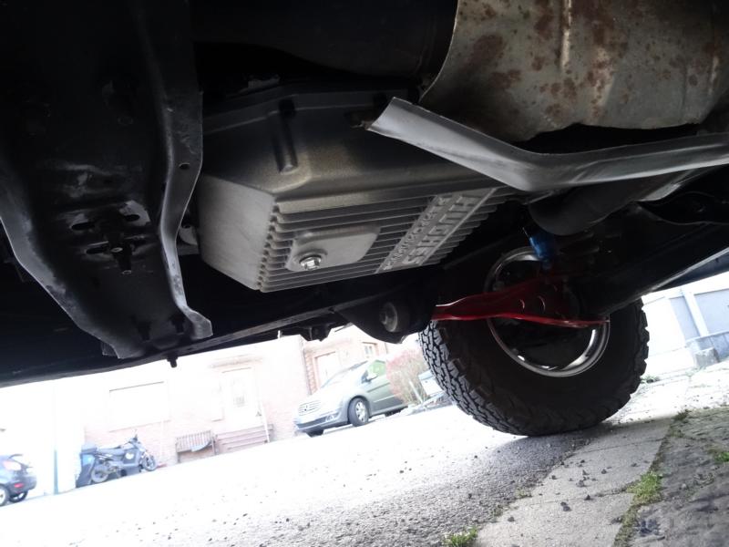 Underbody view of a vehicle showing an aftermarket transmission pan and suspension components. A car and motorcycle are visible in the background
