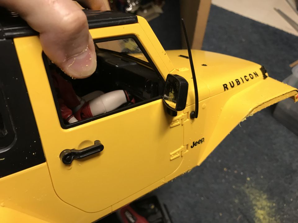 Yellow Jeep Wrangler Rubicon model, side view, with 'Jeep' and 'Rubicon' text visible. Hand holding the roof