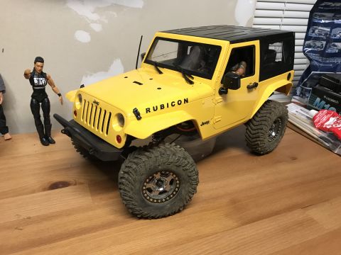 1989 Ford Bronco - A yellow toy Jeep Rubicon with a black top and large off-road tires sits on a wooden surface next to a doll.