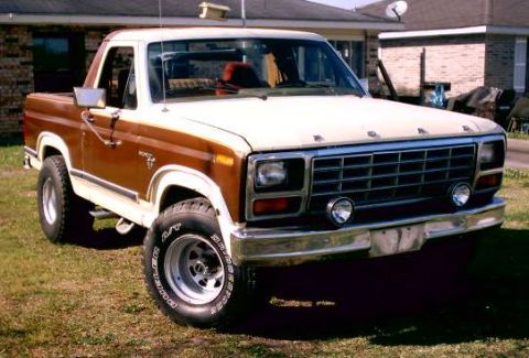 1980 Ford Bronco - Early-1980s Ford F-Series pickup, two-tone brown over tan, chrome bumper and alloy wheels, parked on a grassy yard by a brick house.