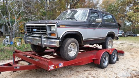 1986 Ford Bronco - A two-tone Ford Bronco is loaded onto a red trailer, ready for transport.