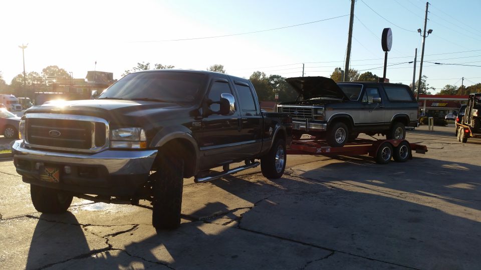 Ford Super Duty pickup towing a full-size Ford Bronco with hood up on a red trailer at a gas station