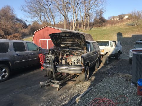 1992 Ford Bronco - A black Ford Bronco with its hood open is parked in a driveway next to a gray SUV. The Bronco is on an engine hoist, suggesting mechanical…