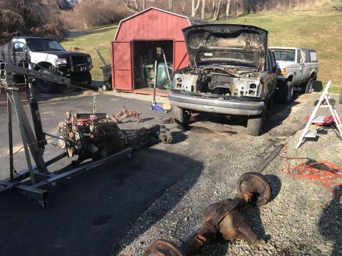 1992 Ford Bronco - A disassembled engine and transmission are suspended from an engine hoist in a driveway next to a Ford truck with its hood open. A Ford Bro…