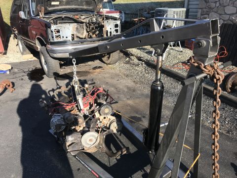 1992 Ford Bronco - A dark-colored Ford Bronco is parked on a gravel and asphalt surface with its hood open, revealing the engine bay. An engine is suspended b…