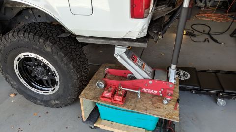 1995 Ford Bronco - A white Ford Bronco is jacked up on a metal plate, with a red Arcan hydraulic jack supporting it from underneath. A set of Milwaukee drill…