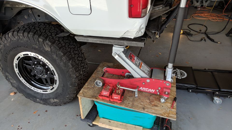 White Ford Bronco lifted by a red hydraulic jack, with drill bits on a wooden platform.