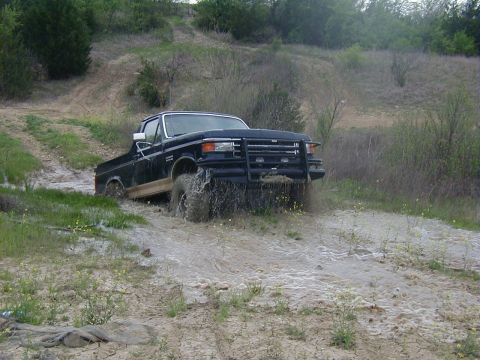 1991 Ford Bronco - A dark blue full-size Ford Bronco with the rear top removed churns through a shallow muddy water crossing at the bottom of an eroded trail,…
