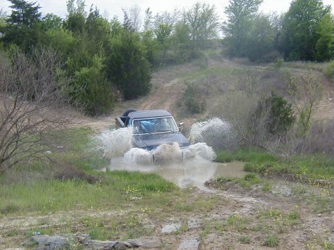 1991 Ford Bronco - Wide shot of a topless dark-colored full-size Ford 4x4 blasting through a shallow water crossing, throwing a heavy wall of spray off both s…
