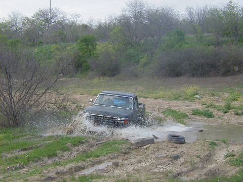 1991 Ford Bronco - Three-quarter front view of a topless dark blue Ford Bronco splashing through a shallow water crossing, spray fanning off the front bumper…