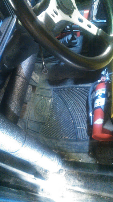 1978 Ford Bronco - The interior of a vehicle shows a black steering wheel, a red fire extinguisher mounted to the floor, and a Ford-branded rubber floor mat.
