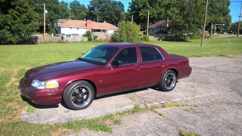 2004 Ford Crown Victoria - A dark red sedan is parked on a cracked asphalt lot next to a grassy area. The car has tinted windows and aftermarket black and chrome whee…