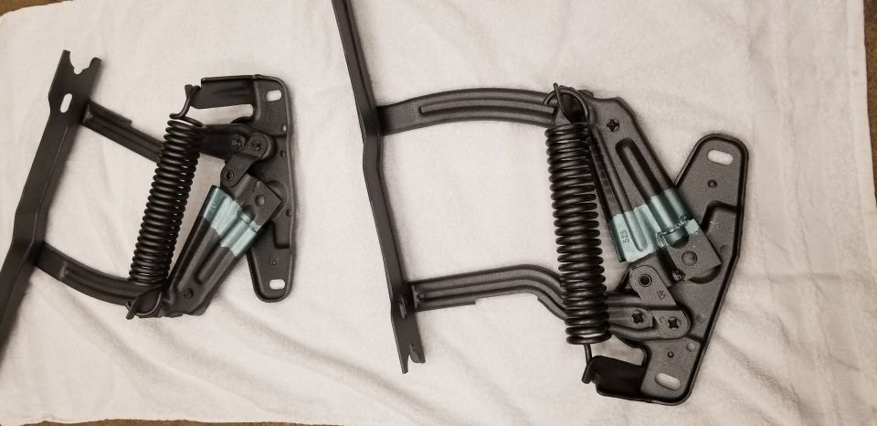 Two black metal hood hinges with springs on a white cloth.