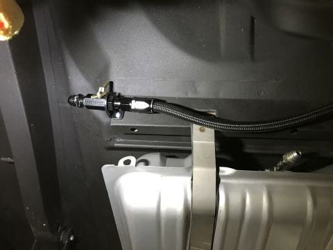 1964 Ford Fairlane - A black braided fuel line with a black fitting and a lever shut-off valve is mounted to the undercarriage of a vehicle, near a silver fuel…