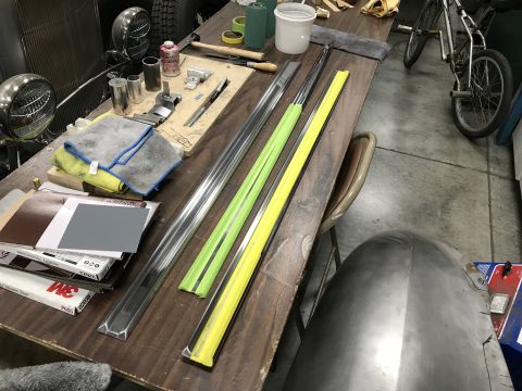 1964 Ford Fairlane - A workbench is laid out with various automotive parts and tools, including long metal trim pieces, sandpaper, and microfiber cloths. A port…