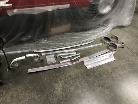 1964 Ford Fairlane - Various polished aluminum trim pieces and headlight bezels are laid out on a concrete floor next to a red vehicle covered in plastic sheeti…